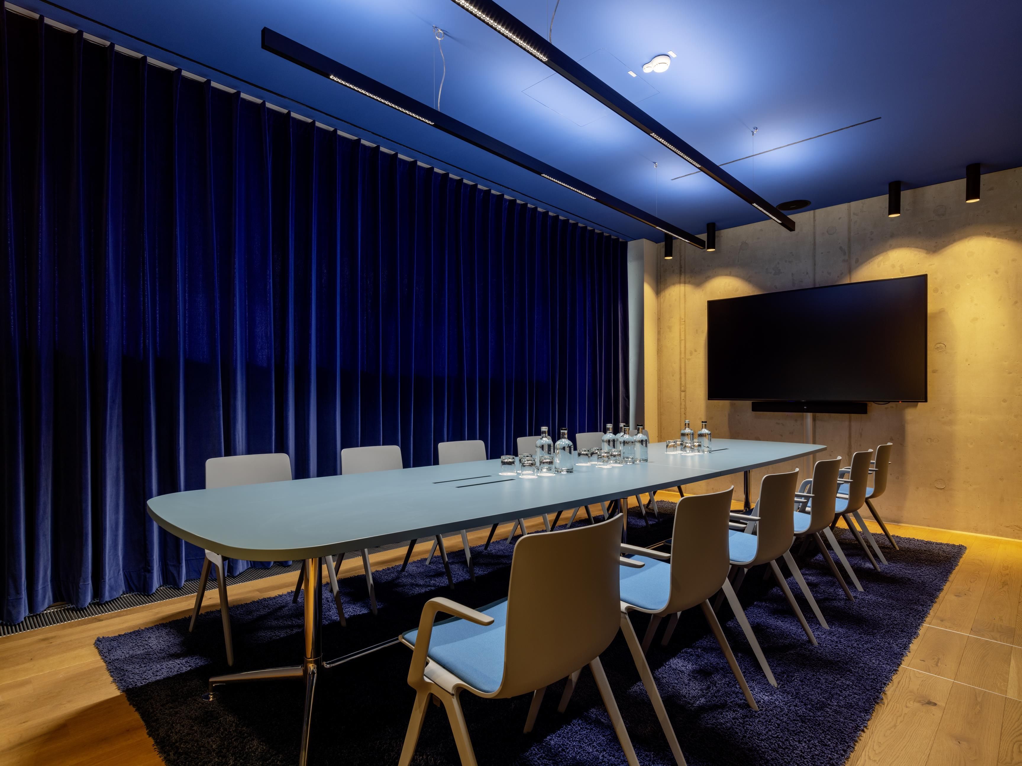 A conference room with a long table, chairs, a flat-screen TV, and a blue curtain backdrop.