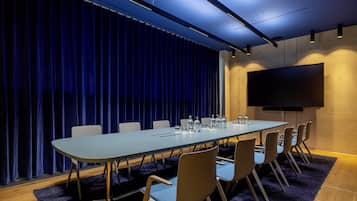 A conference room with a long table, chairs, a flat-screen TV, and a blue curtain backdrop.