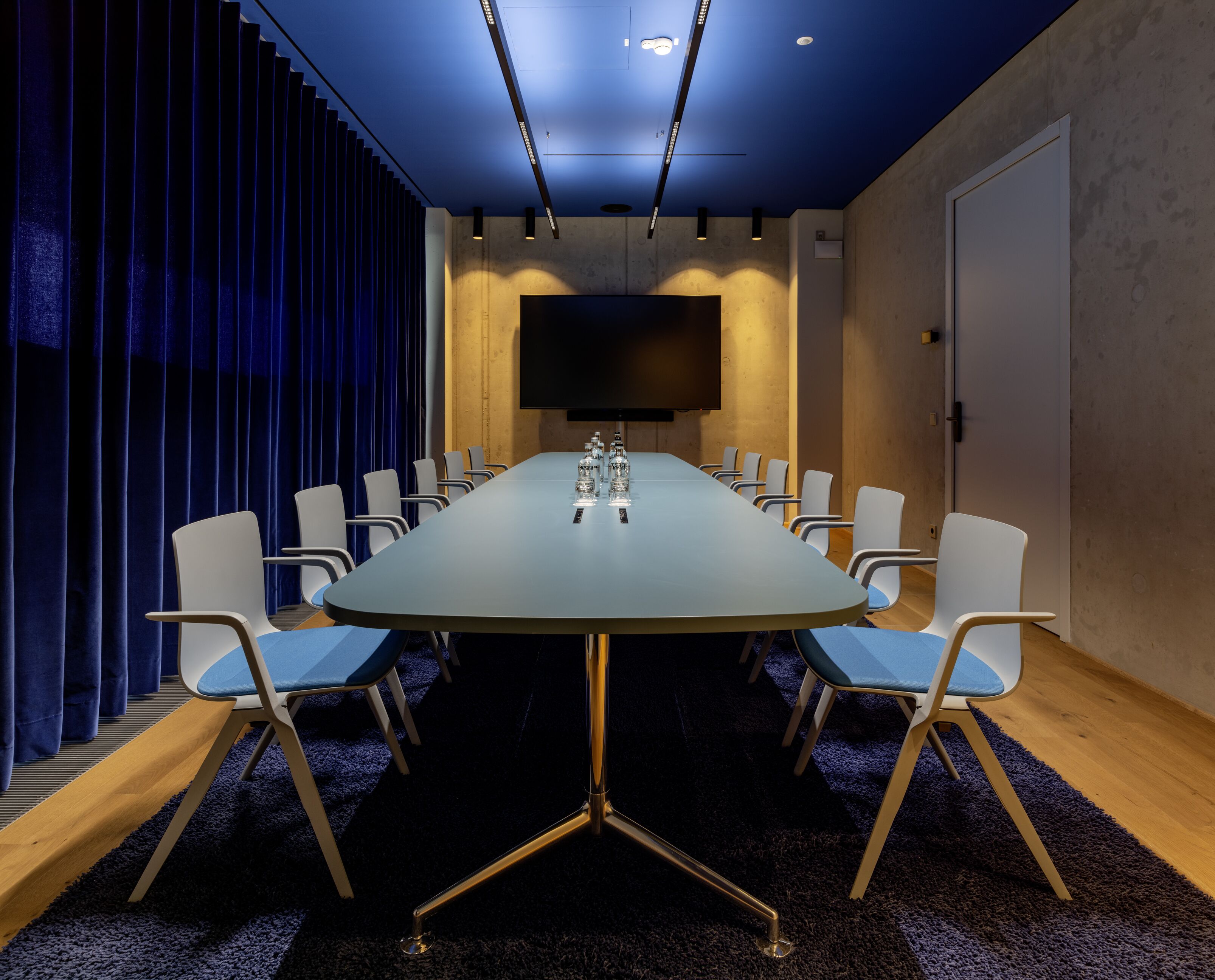 Une salle de conférence avec une longue table, des chaises, un téléviseur à écran plat et un fond bleu en forme de rideau.