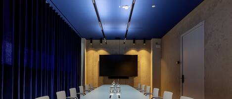 A conference room with a long table, chairs, a flat-screen TV, and a blue curtain backdrop.