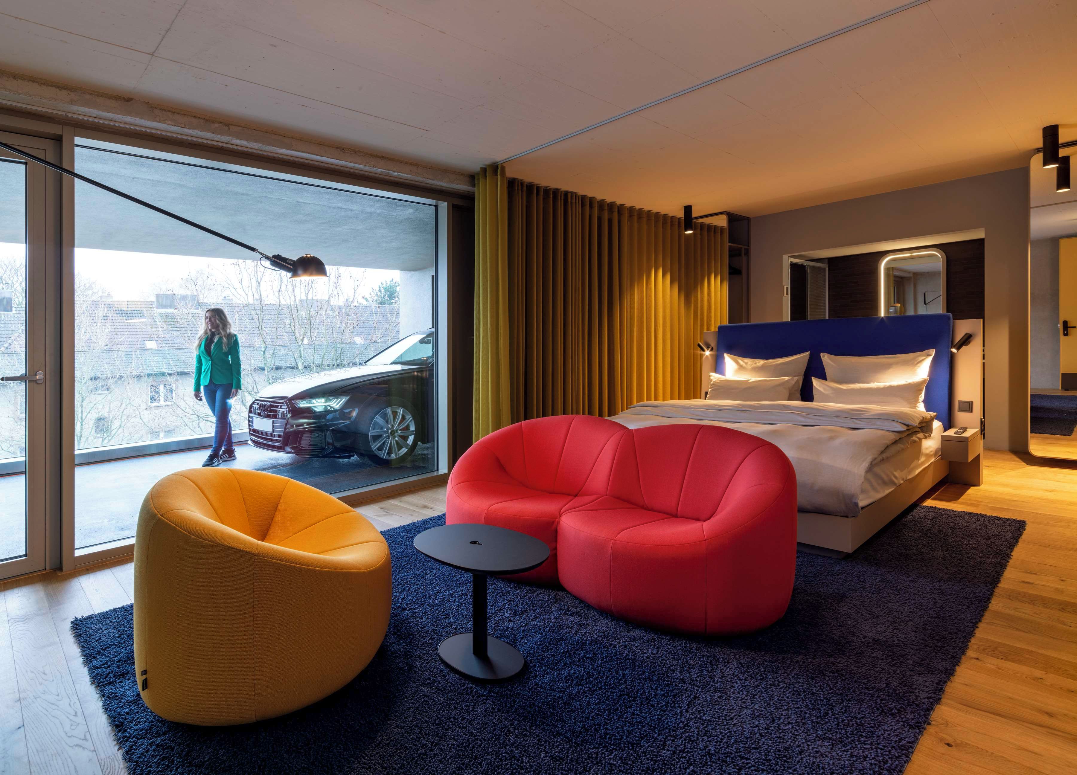 A modern hotel room with a large bed, a red armchair, a yellow bean bag chair, a small round table, and a large window with a view of a parked car and buildings outside.