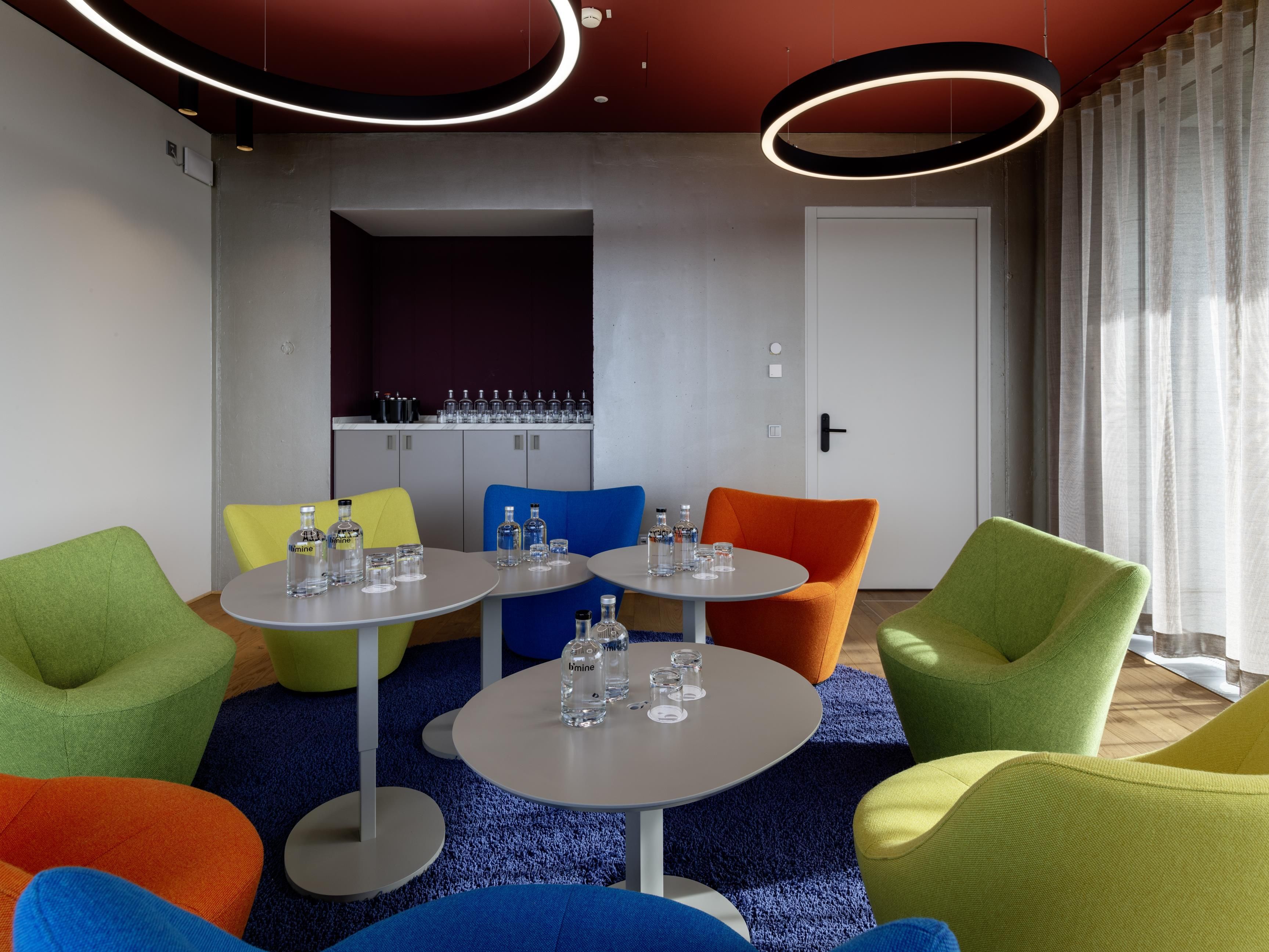 A modern hotel room with a seating area featuring colorful chairs, a small table with glasses, and a minibar.