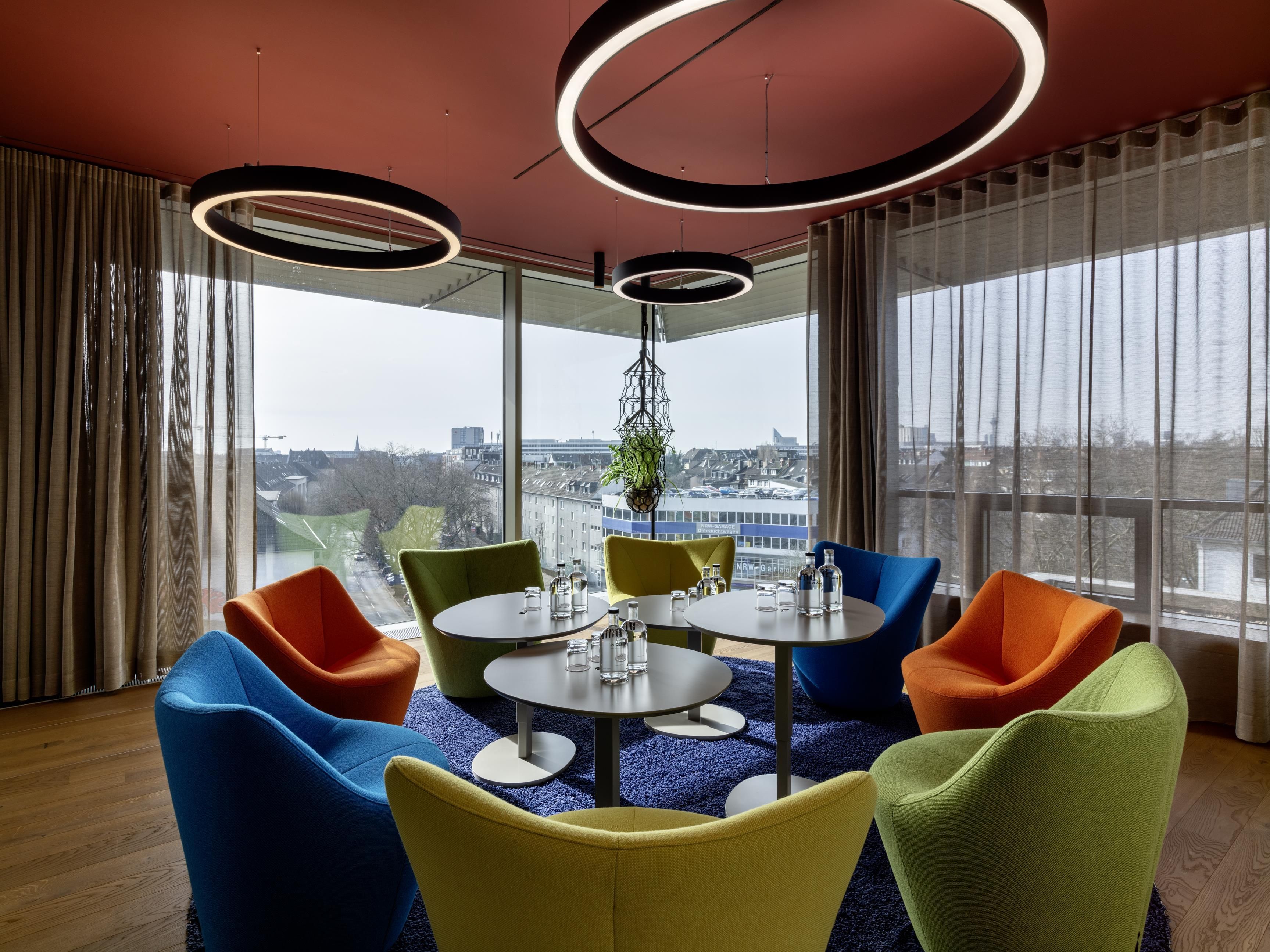 A modern meeting room with a round table, colorful chairs, and large windows offering a city view.