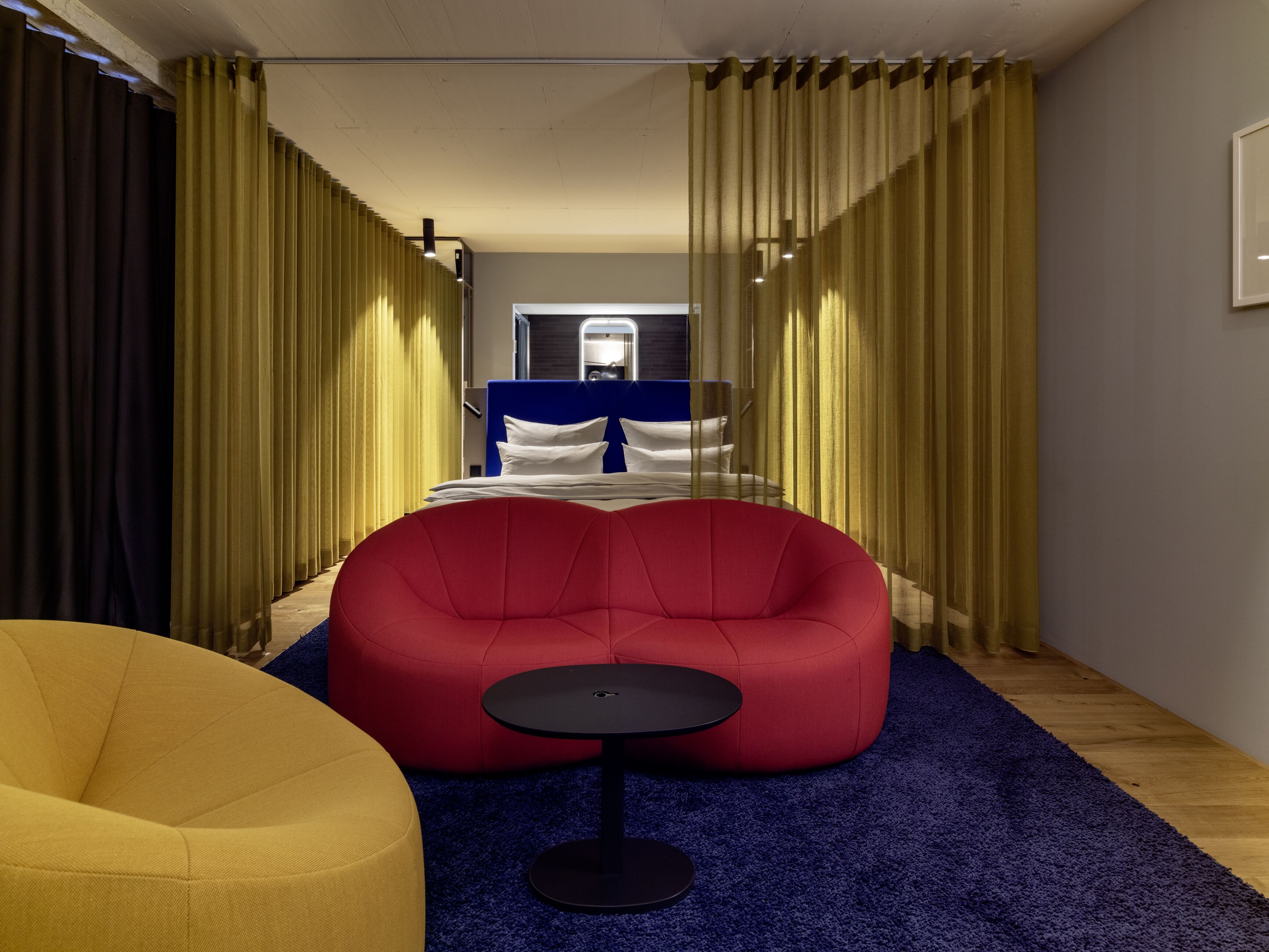 Een moderne hotelkamer met een groot bed, een rode relaxfauteuil, een klein rond tafeltje en een blauw vloerkleed.