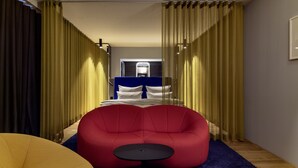 Une chambre d’hôtel moderne avec un grand lit, un fauteuil en forme de poire rouge, une petite table ronde et une moquette bleue.