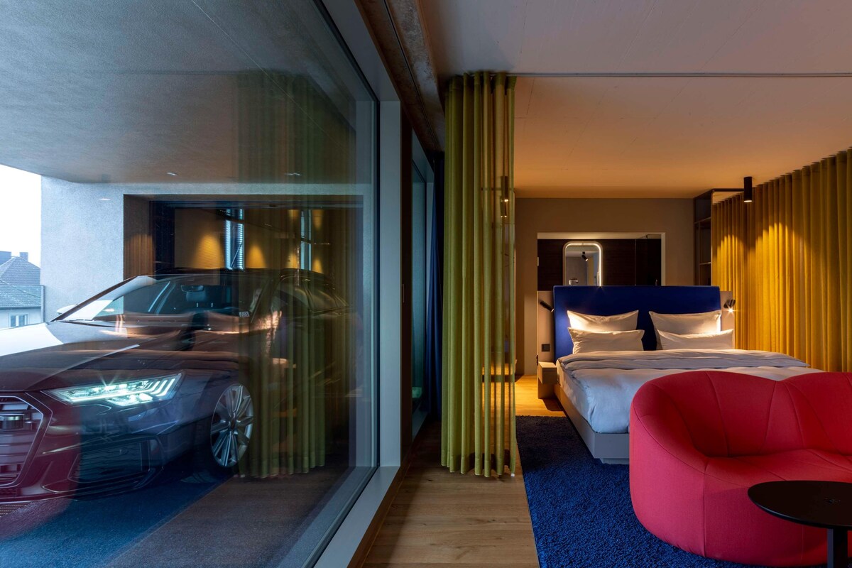 Une chambre d’hôtel moderne avec une grande fenêtre, un lit, un fauteuil rouge et une voiture garée à l’extérieur.