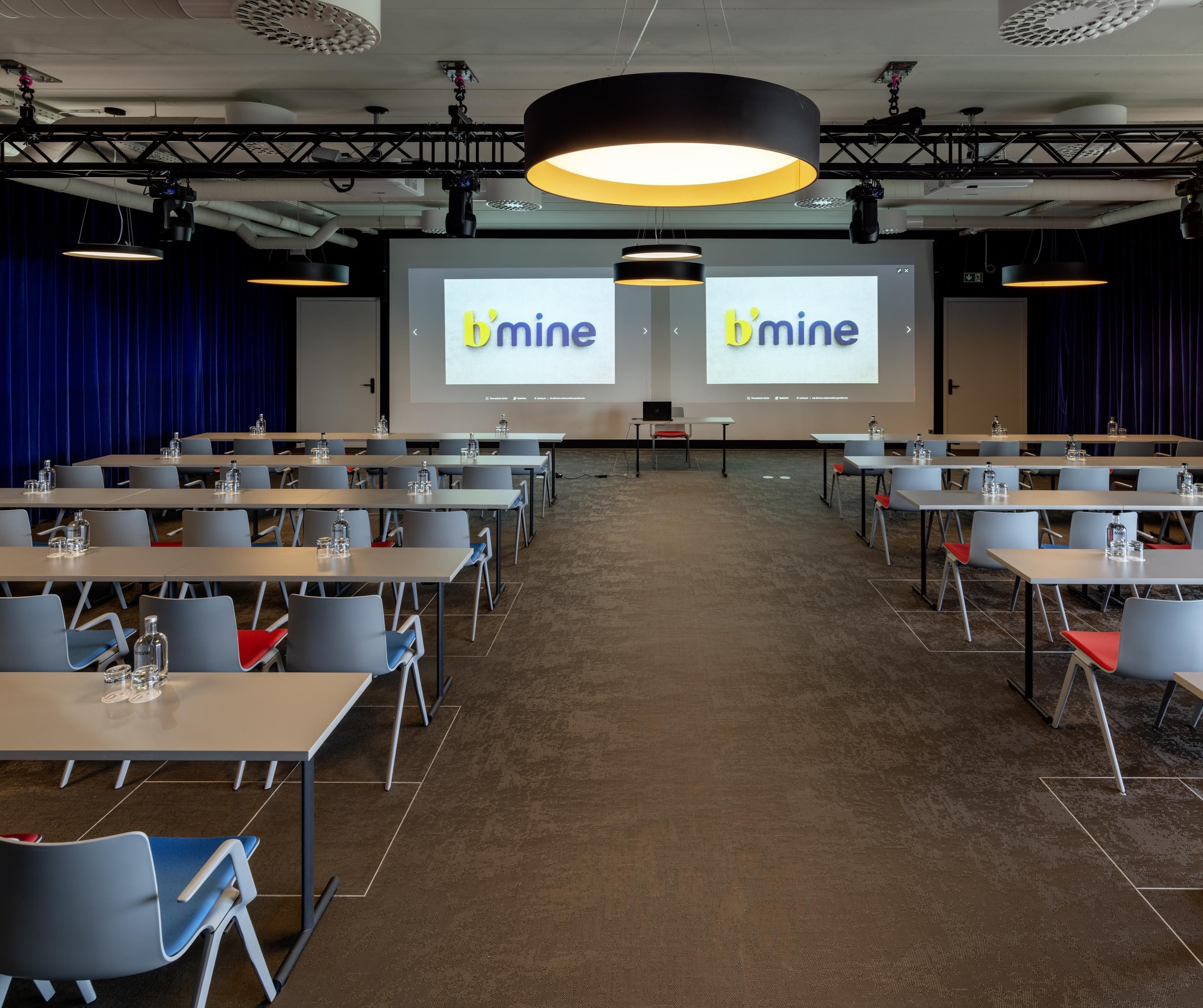 A conference room with multiple tables and chairs, a large screen displaying the 'b'mine' logo, and overhead lighting.