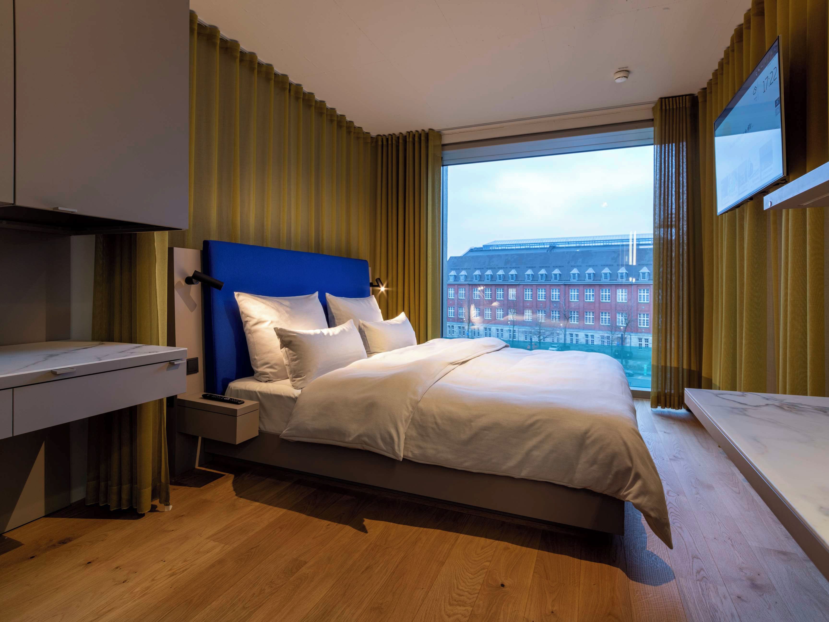 Ein modernes Hotelzimmer mit einem großen Bett, einem Schreibtisch und Blick auf eine Stadtlandschaft durch ein großes Fenster.
