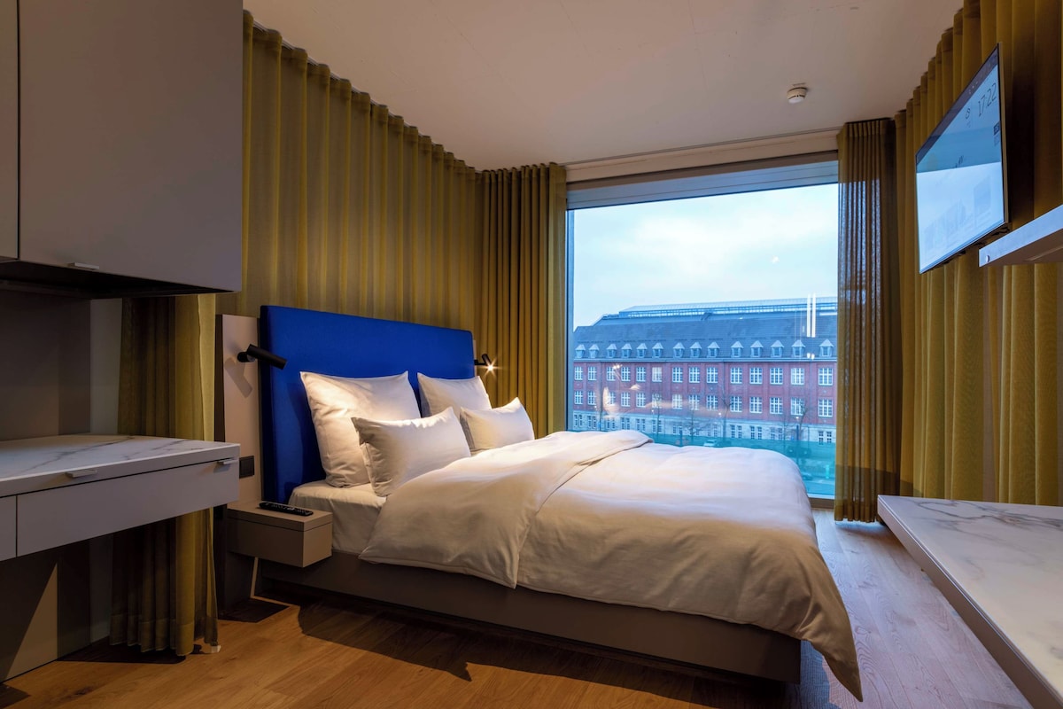 Una camera d'albergo moderna con un letto grande, una scrivania e vista sulla città attraverso un'ampia finestra.