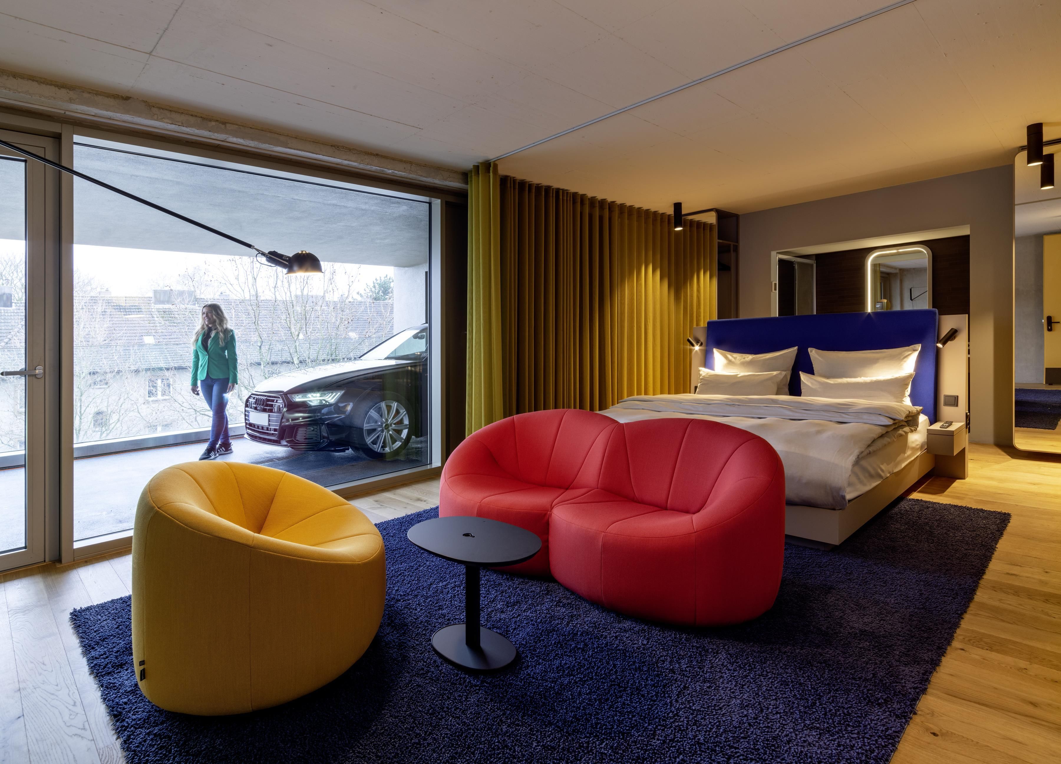 A modern hotel room with a large sliding glass door, a bed with a blue headboard, a red armchair, a yellow bean bag chair, and a small round table.