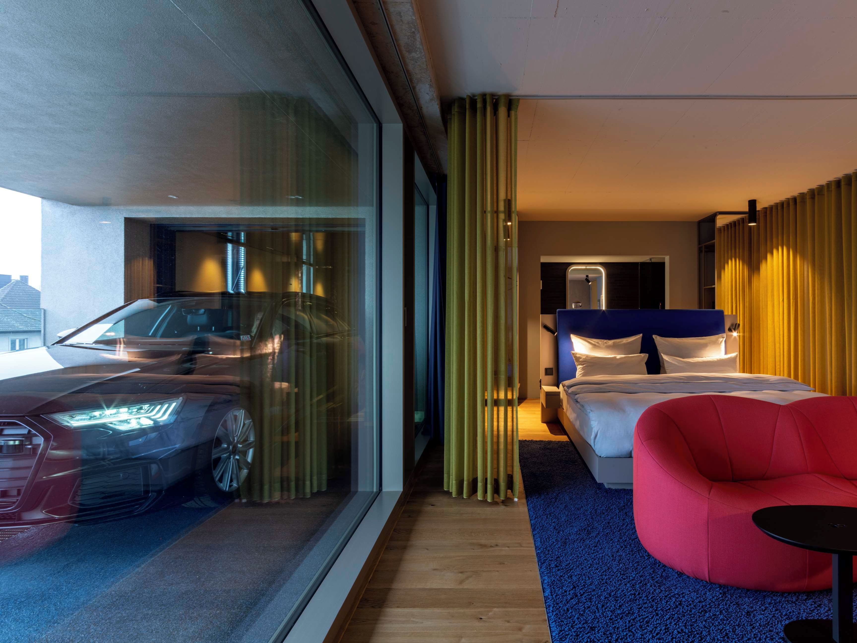 Une chambre d’hôtel moderne avec une grande fenêtre, un lit doté d’une tête de lit bleue, un fauteuil rouge et une voiture garée à l’extérieur.