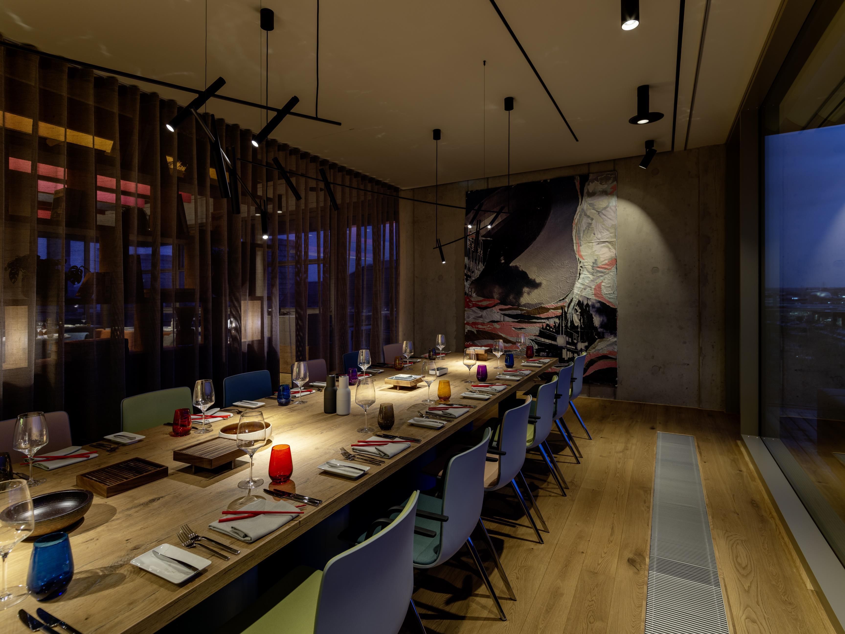 A long dining table set for a meal with chairs, a cityscape mural on the wall, and a view of the city through large windows.