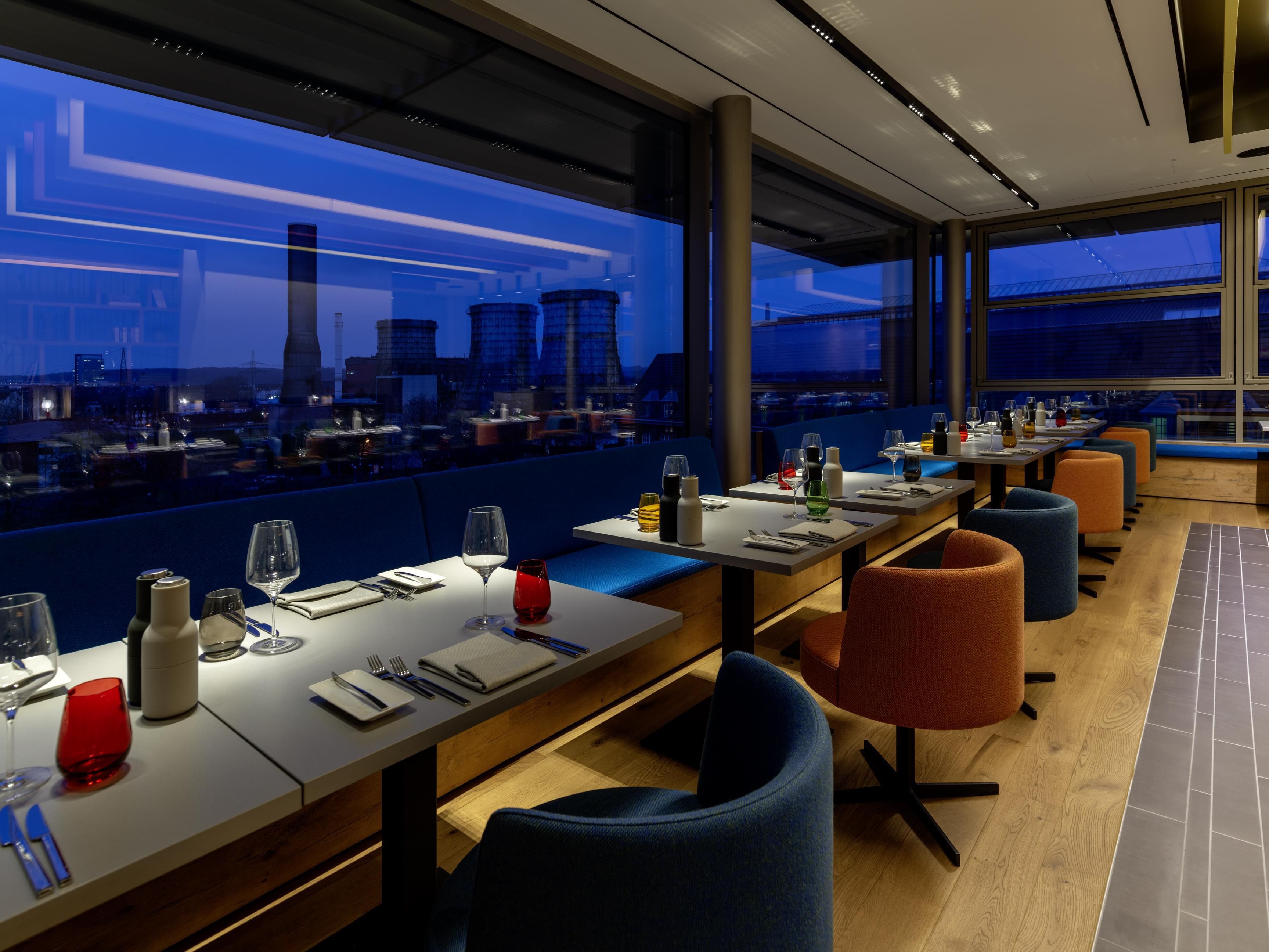 A restaurant with tables set for dining, large windows offering a city view, and modern seating.