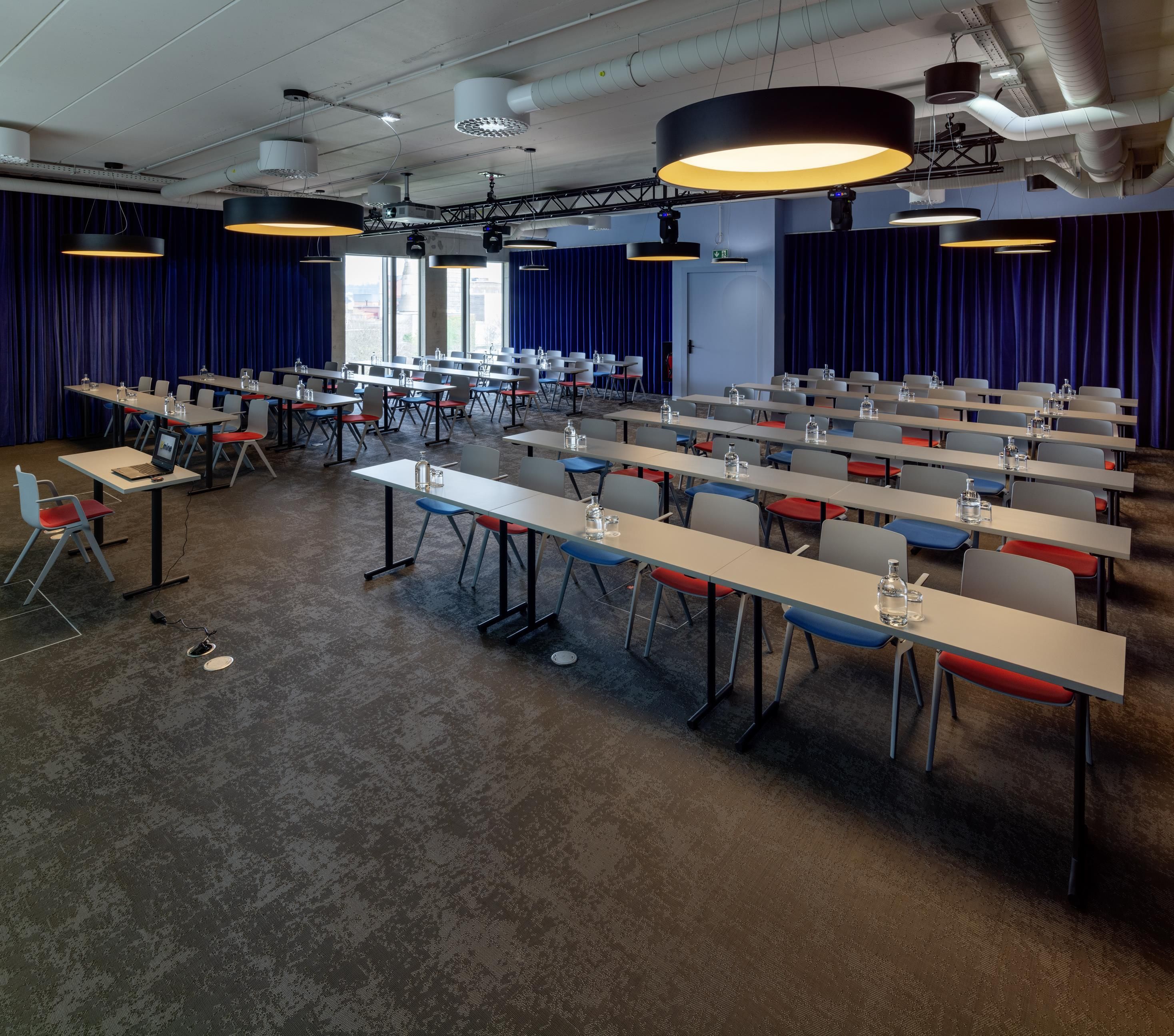 Een vergaderruimte met rijen tafels en stoelen, grote hanglampen en een stadszicht door grote ramen.