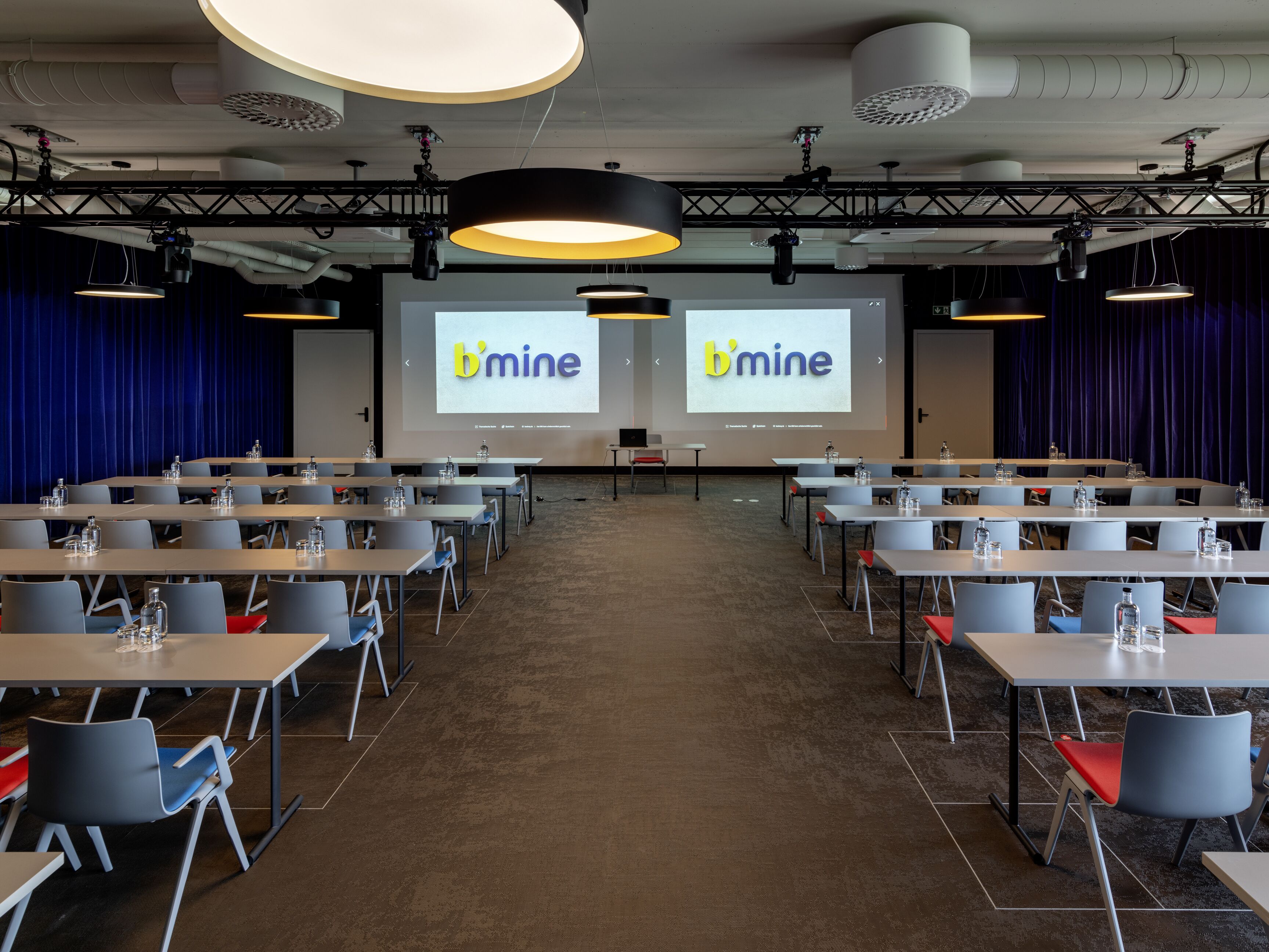 Un salón de conferencias con varias mesas y sillas, una pantalla grande mostrando el texto 'b'mine' y un escenario con equipo de iluminación.