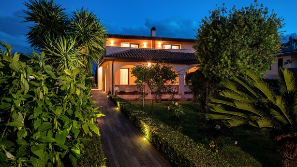 Una casa bien iluminada con techo de tejas, rodeada de exuberante vegetación y un camino que lleva a la entrada.