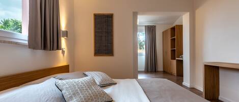 Une chambre d’hôtel moderne avec un grand lit, une tête de lit en bois, une table de chevet, une lampe fixée au mur et une vue sur la salle de bain, accessible par une porte ouverte.