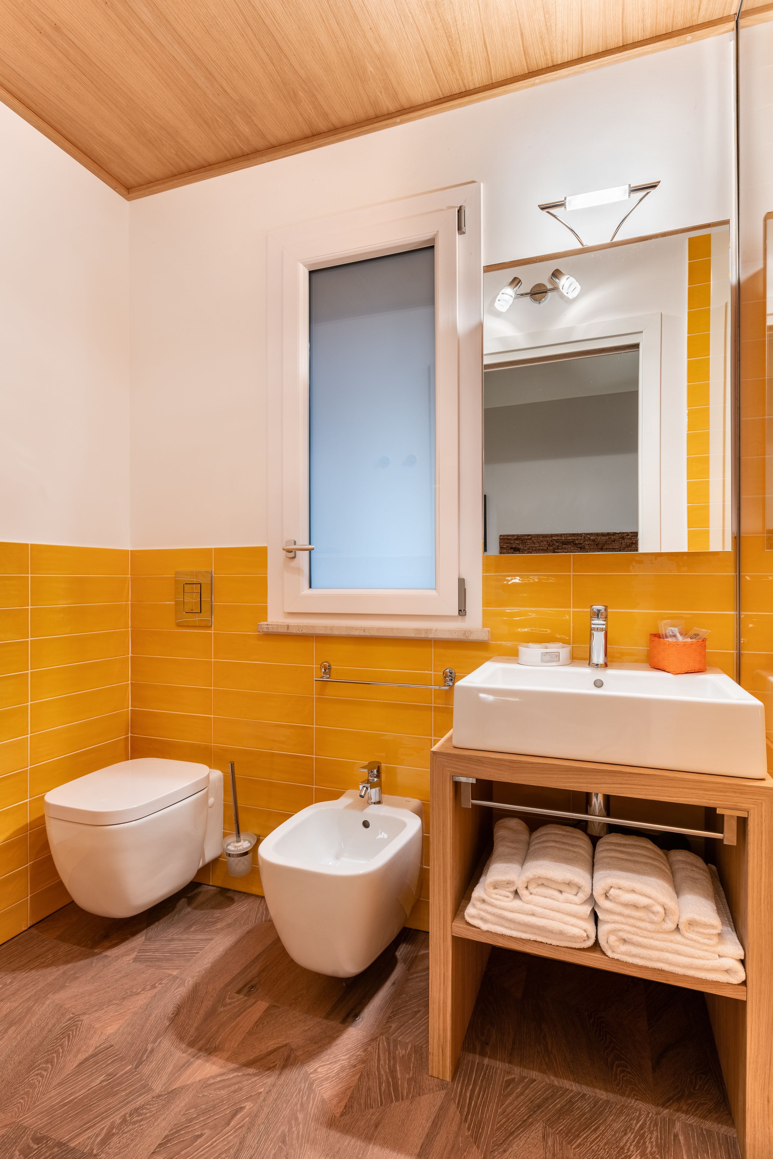 A bathroom with a white toilet, a bidet, a sink with a mirror above, and a shelf with folded towels.
