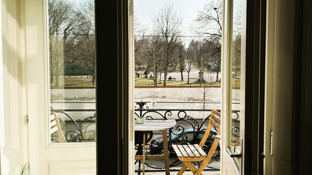 Vue depuis une chambre avec balcon, comprenant une table et des chaises, une balustrade et une vue sur un parc avec des arbres et des bancs.