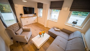Una sala de estar moderna con un sofá, un sillón y una mesa de centro. Hay un televisor de pantalla plana montado en la pared, y la habitación tiene ventanas grandes con cortinas.