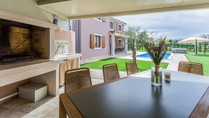 Une terrasse extérieure avec une table à manger, des chaises, un barbecue et une vue sur une cour arrière avec une piscine et un jardin.