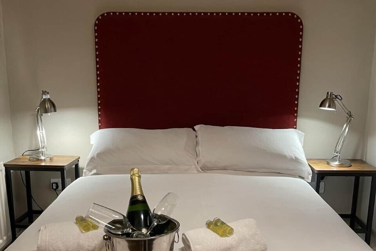 A hotel room with a large bed, a red headboard, two bedside tables with lamps, and a minibar with a bottle of champagne and glasses.