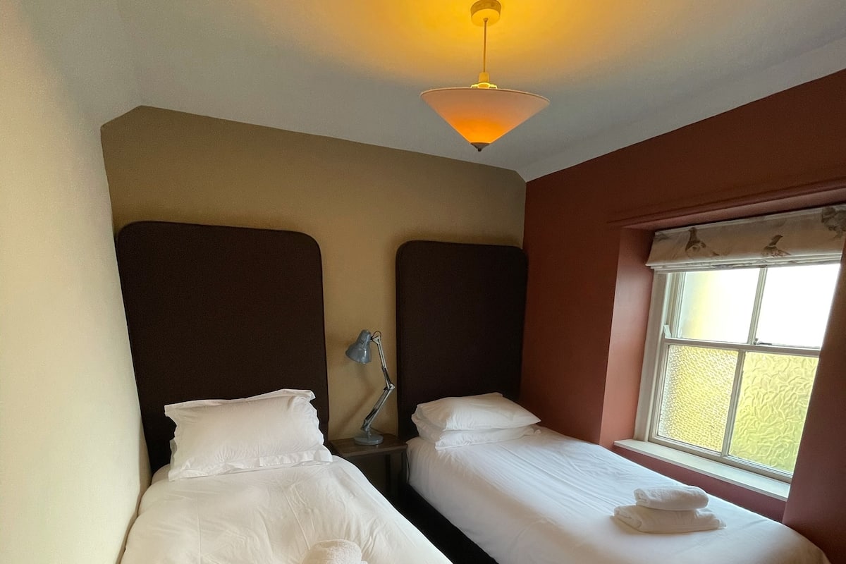 A hotel room with two single beds, a desk with a lamp, a window with a view, and a ceiling light.