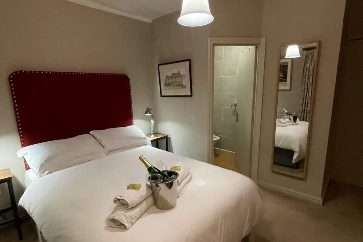 A hotel room with a large bed, a red headboard, a bedside table with a lamp, a framed picture on the wall, and a mirror reflecting the room.