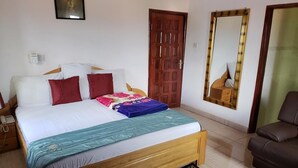 A bedroom with a wooden bed, red and white pillows, a blue and teal blanket, a giraffe-patterned rug, a brown leather chair, and a wooden door. - Nabegyi Hotel (North Legon)