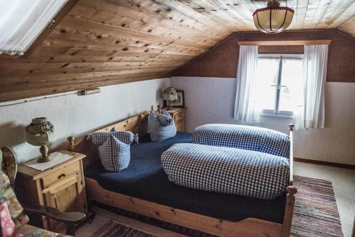 Une chambre mansardée chaleureuse avec un plafond en bois, un lit simple avec une literie à carreaux, une table de chevet, une lampe et une fenêtre avec des rideaux.