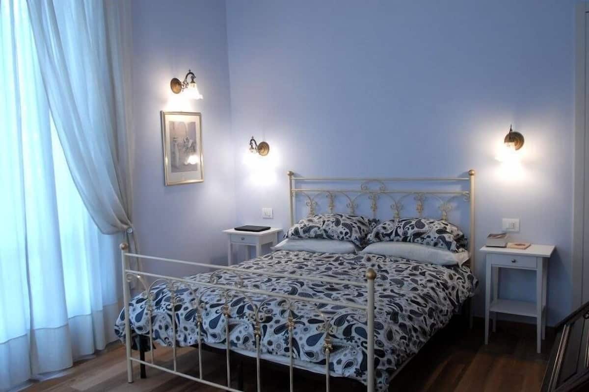 A bedroom with a metal bed frame, patterned bedding, bedside tables, and wall-mounted lights.