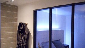 A bathroom with a bathtub, a towel rack with a robe, and a window with a view of a bed and a nightstand.
