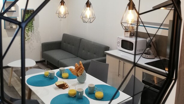 A modern kitchen with a dining area, featuring a geometric ceiling design, pendant lighting, and a grey sofa.