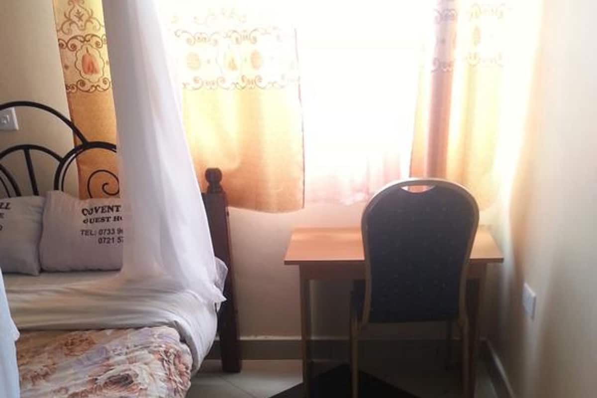Un dormitorio con cama, escritorio y silla. Hay una ventana con cortinas, una mesita de noche con lámpara y una pared con un diseño decorativo.