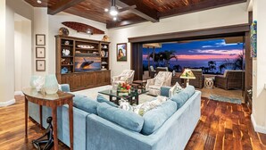 A spacious living room with a large sofa, a coffee table, and a television. There is a wooden ceiling and a view of the ocean through large windows. - 🏖️Ocean View Kona Home | Saltwater Pool + Private Suites (Kailua Kona)