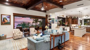 A spacious living room with a large sofa, a coffee table, and a view of palm trees through a sliding glass door. - 🏖️Ocean View Kona Home | Saltwater Pool + Private Suites (Kailua Kona)