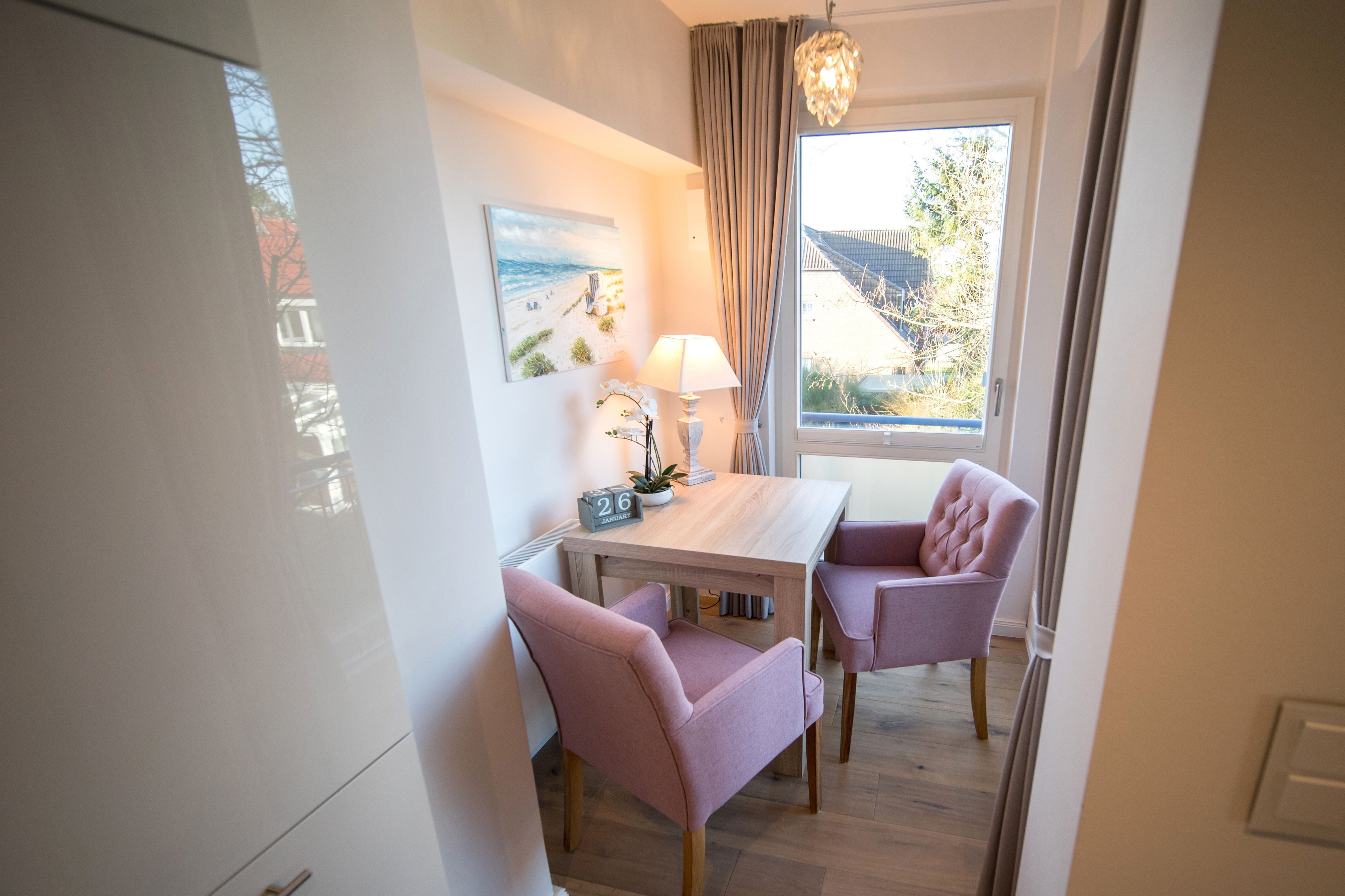 A small dining area with a wooden table, two pink chairs, a lamp, and a window with curtains.