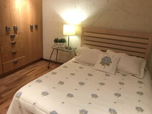 Relax in a serene space with a comfortable bed, a wooden wardrobe, and a small table with a lamp and potted plant.