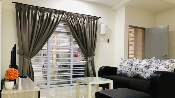 A modern living room with a black sofa, a white coffee table, and a black ottoman. There is a window with curtains, a small table with a vase of flowers, and a white wall-mounted lamp. - De Little Cabin near Sunway Lost World of Tambun Ipoh, Perak Malaysia. (Ipoh)