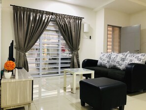 Un salon moderne avec un canapé noir, une table basse blanche et un pouf noir. On y trouve une fenêtre avec des rideaux, une petite table sur laquelle est posé un vase de fleurs et une lampe murale blanche.