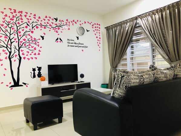 Un salon doté d’un téléviseur à écran plat, d’un canapé noir, d’un petit pouf et d’un mur décoratif représentant un arbre et des oiseaux.