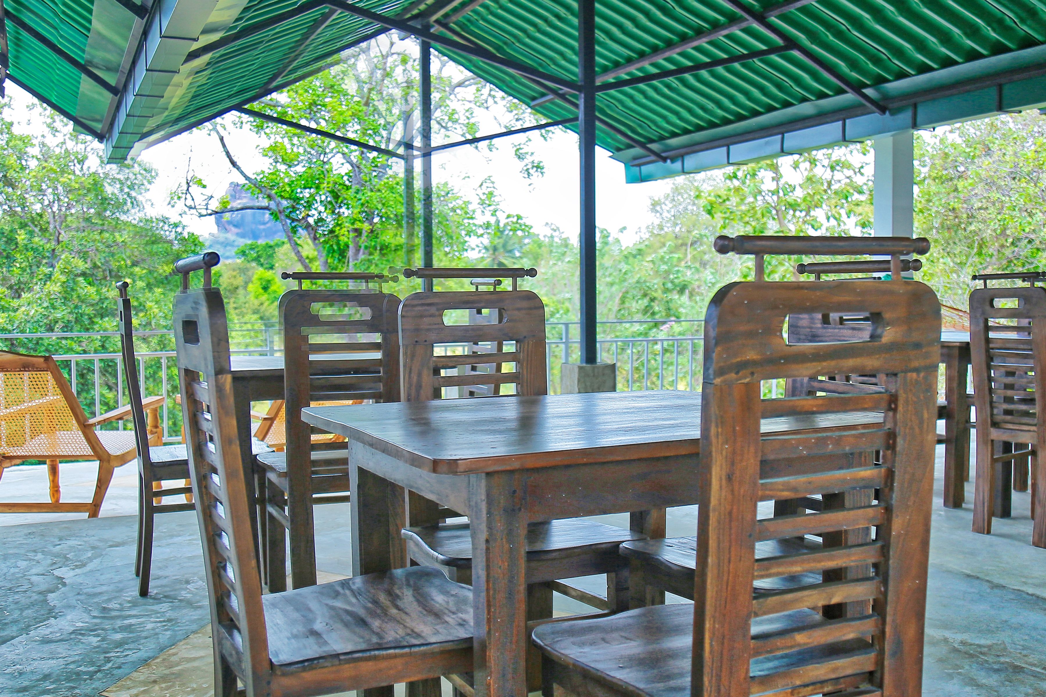 A covered outdoor seating area with wooden tables and chairs, surrounded by greenery.