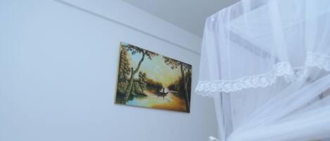 A room with a white wall, a painting of a river scene, and a white canopy bed.