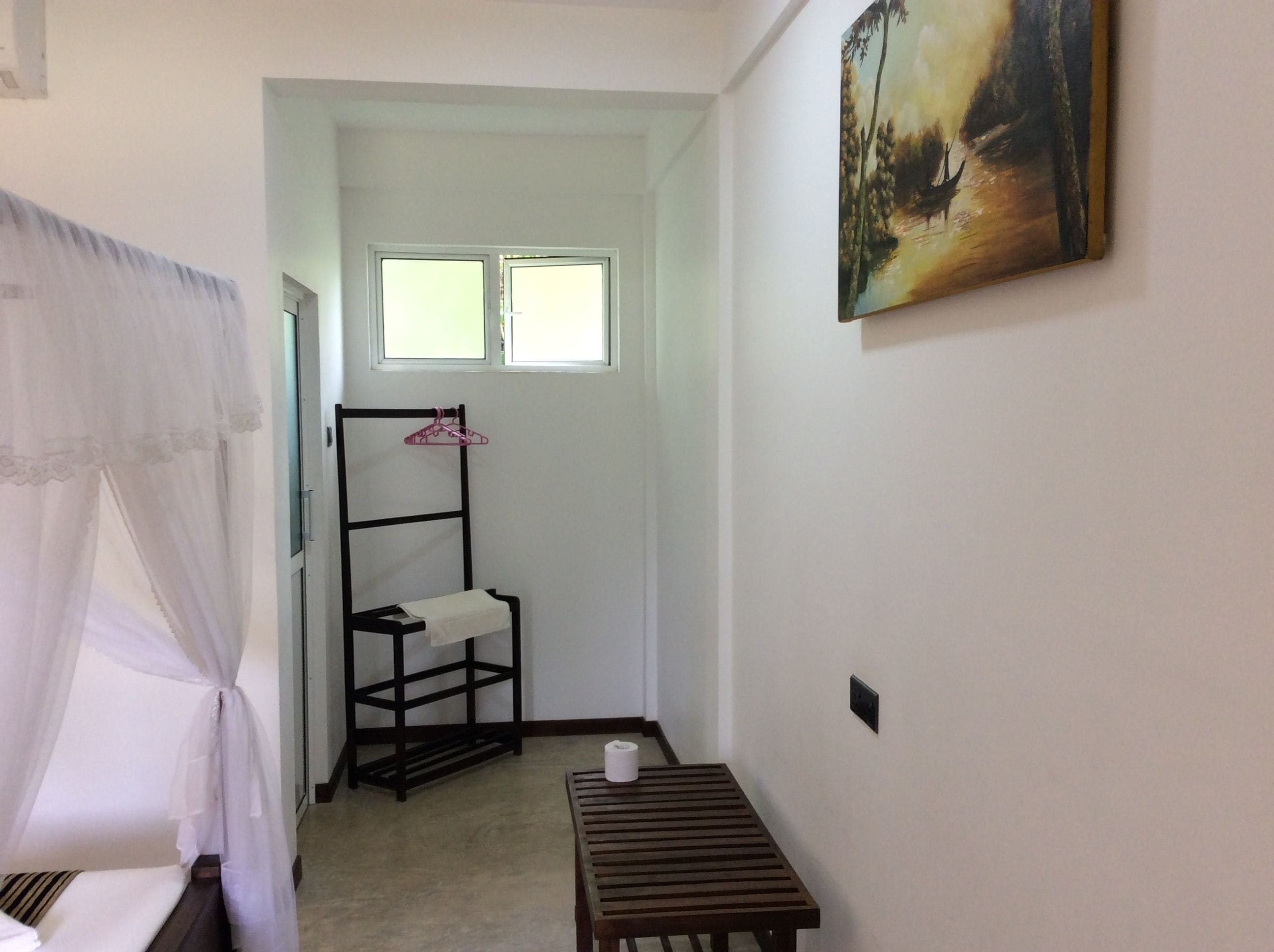 Una habitación pequeña y limpia con una cama, un banco de madera, un perchero y un cuadro en la pared.