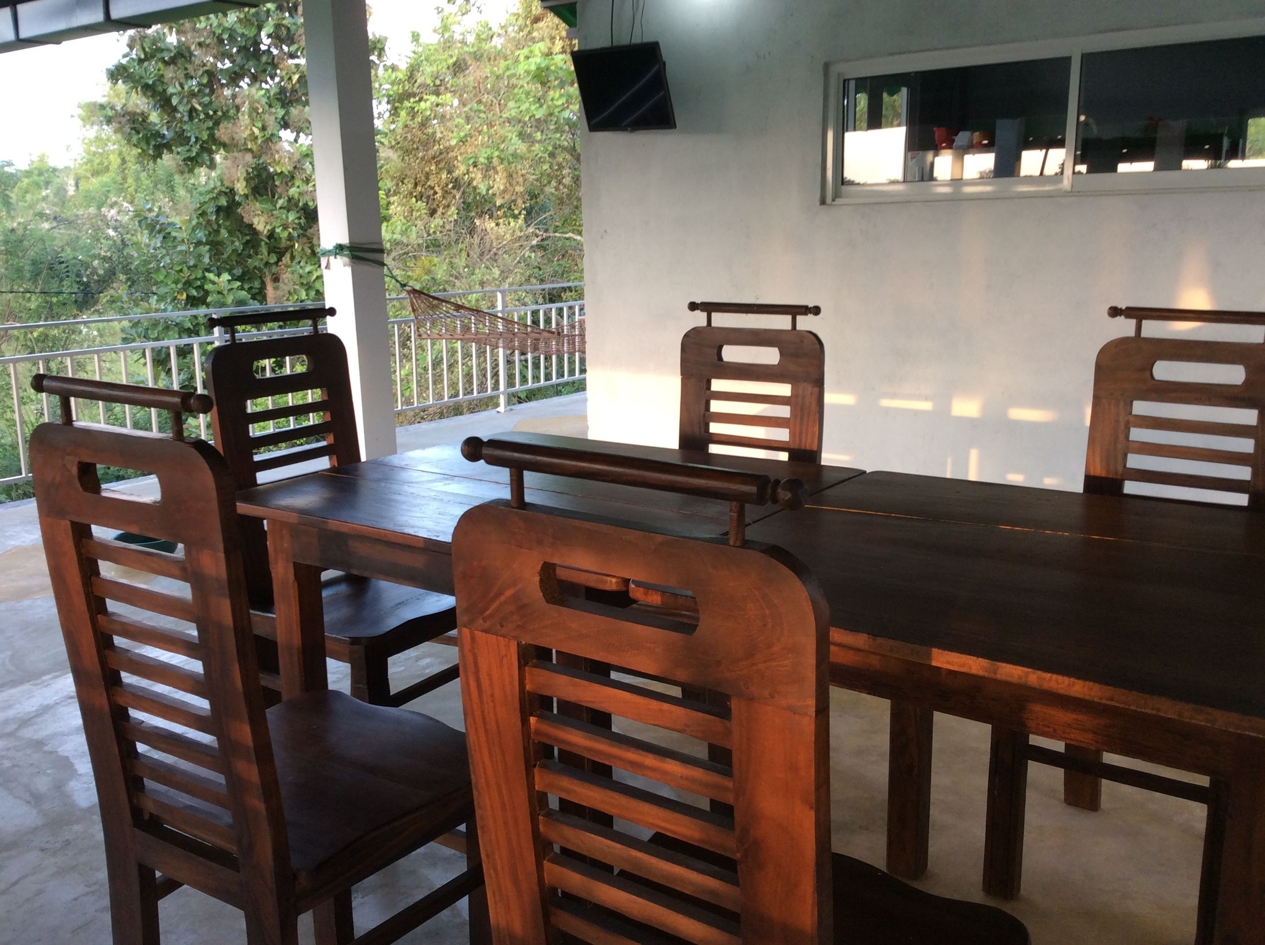 A wooden dining table with chairs, a TV mounted on the wall, and a view of greenery outside.