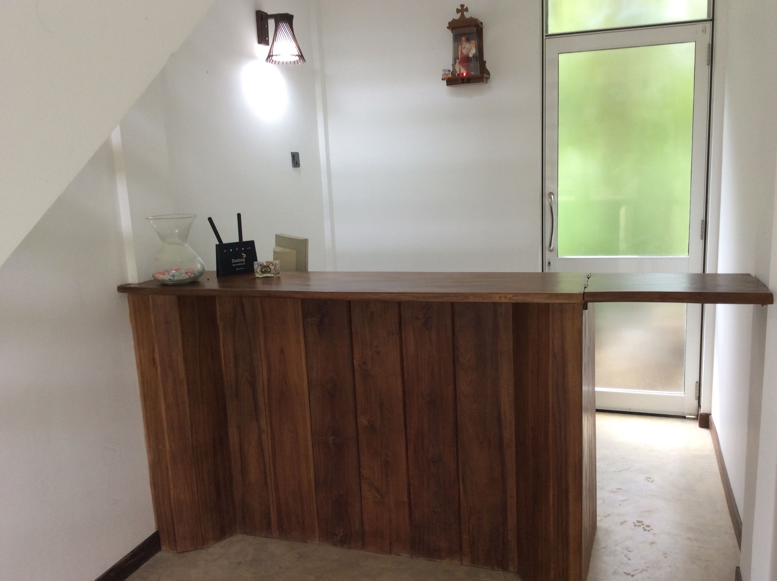 Un comptoir d’accueil en bois, situé dans un couloir, avec un luminaire fixé au mur et une petite statue religieuse.