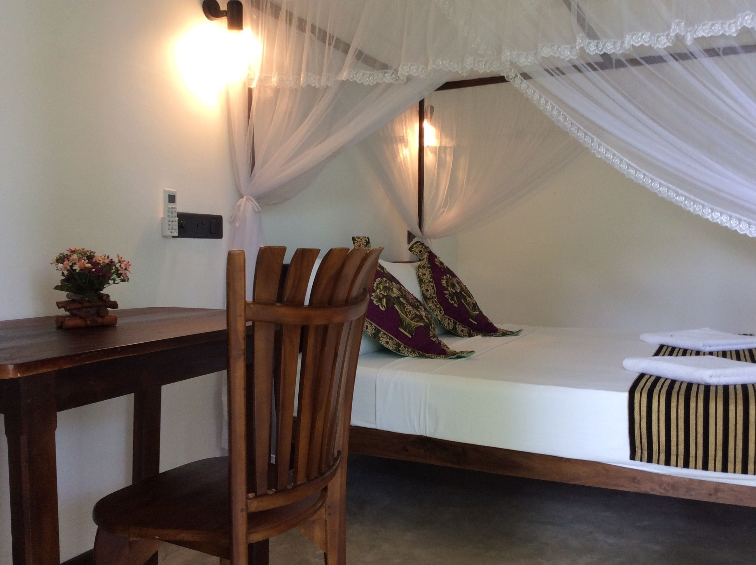 Una habitación con una cama bajo un dosel, una silla de madera, una mesita de noche y una lámpara fijada a la pared.