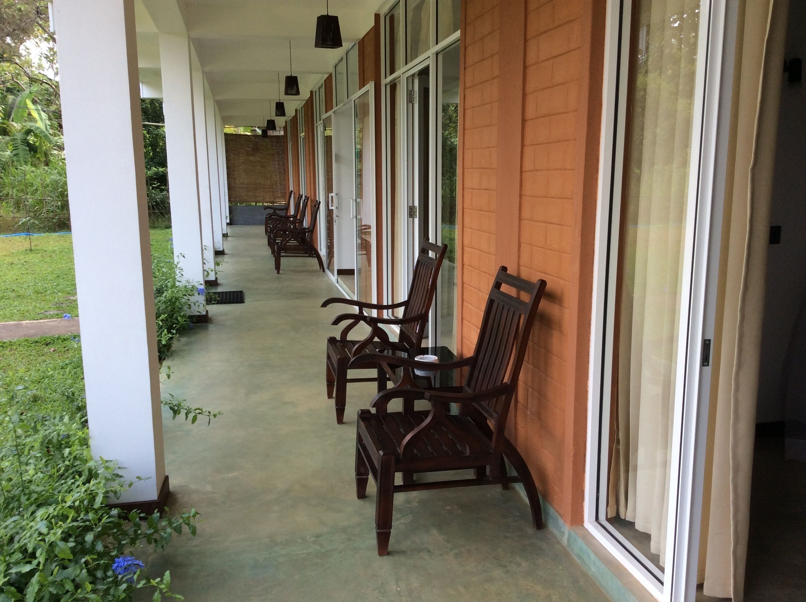 A covered outdoor seating area with wooden chairs and tables, a brick wall, and large windows.