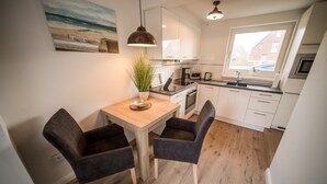 Una cocina compacta con armarios blancos, una mesa de comedor de madera, dos sillas, una lámpara colgante y una planta en maceta.