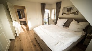 Un dormitorio con una cama grande, suelo de madera, una puerta que da a otra habitación y una ventana con cortinas.